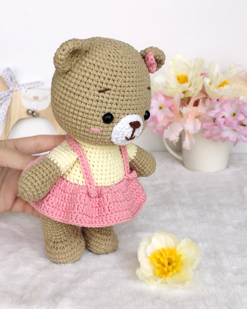 May include: A crocheted brown bear wearing a pink and yellow dress. The bear has black eyes and a white nose. The bear is standing on a white fluffy surface.