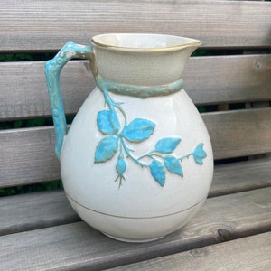 May include: A white ceramic pitcher with a blue floral design. The pitcher has a handle and a wide mouth.