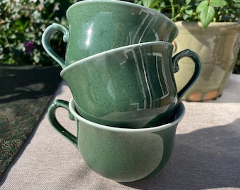 May include: Three stacked green ceramic cups with handles. The cups have a speckled texture and a white rim. The background includes green foliage and a pot with pink and red roses.