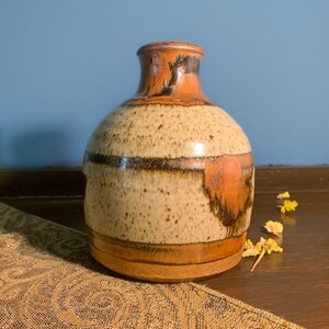 May include: A small, brown and tan ceramic vase with a wide mouth and a narrow neck. The vase has a speckled glaze with brown and orange stripes.