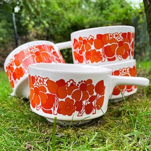 Könnte beinhalten: Vier weiße Keramikbecher mit einem floralen Muster in Orange- und Weißtönen. Die Becher haben eine abgerundete Form und einen Henkel.
