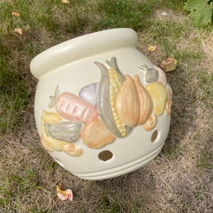 Puede incluir: Recipiente para ajos de cerámica color crema con verduras en relieve. El recipiente presenta una variedad de verduras coloridas, incluyendo zanahorias, maíz y ajo. Tiene tres agujeros redondos cerca de la parte inferior y una abertura ancha en la parte superior.