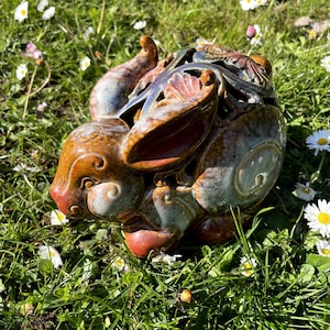 Könnte beinhalten: Eine Keramik-Kaninchenfigur mit detaillierter, glasierter Oberfläche. Das Kaninchen ist hauptsächlich braun mit Akzenten in Rot, Blau und Grün. Die Figur steht in grünem Gras mit kleinen weißen Blumen.