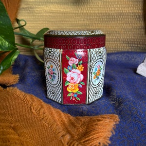 Puede incluir: Un contenedor de hojalata vintage con un diseño floral blanco y rojo. El contenedor tiene una forma hexagonal y una tapa con un patrón decorativo.
