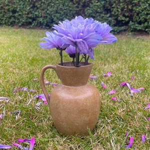 May include: A brown ceramic jug with a wide mouth and a handle, filled with a bouquet of purple flowers. The jug is sitting on a bed of green grass with scattered purple flower petals.