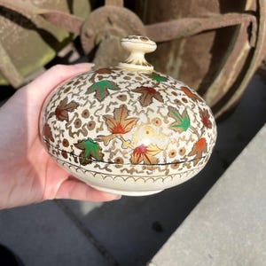 Pode incluir: Uma caixa de bugigangas creme com tampa e design floral. A caixa apresenta um padrão de folhas douradas, vermelhas e verdes. A tampa tem um pequeno botão redondo. A caixa é segurada em uma mão.