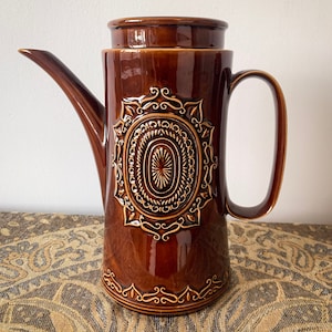 May include: A brown ceramic coffee pot with an ornate design. The pot has a lid and a handle. The design features a large oval with a smaller oval inside, surrounded by a ring of smaller ovals.