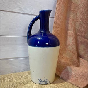 Peut inclure: Une cruche en céramique blanche avec un dessus et une poignée bleus. La cruche porte l'inscription "Dolfi" en bleu sur le fond.