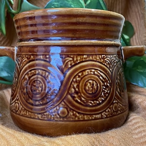 Puede incluir: Una olla de cerámica marrón con dos asas y un patrón decorativo. La olla tiene un acabado brillante y está decorada con un patrón repetitivo de círculos y espirales.