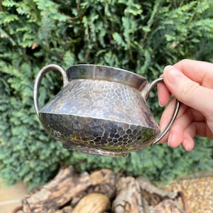 May include: A silver sugar bowl with a hammered finish and two handles. The bowl is slightly tilted and has a reflective surface.