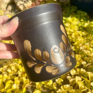 Pode incluir: Uma caneca de cerâmica marrom com um design floral estilizado em uma cor marrom mais clara. A caneca tem uma forma arredondada e uma borda ligeiramente alargada.