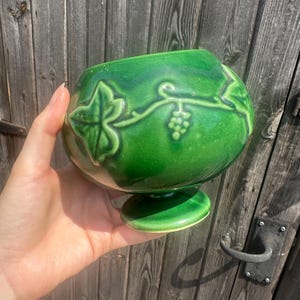 Può includere: Una ciotola in ceramica verde scuro lucido caratterizzata da un motivo in rilievo con foglie di edera e viticci. La ciotola presenta un corpo arrotondato e bulboso che poggia su una piccola base circolare, con una finitura liscia e riflettente.