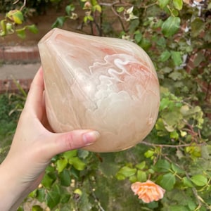 May include: A teardrop-shaped decorative vase with a marbled design in shades of peach, cream, and white. The vase has a wide base and a narrow opening. The background features green foliage and peach-colored roses.