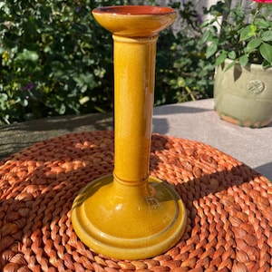 Może przedstawiać: Wysoki, żółty ceramiczny świecznik z szeroką podstawą i rozszerzaną górą. Wnętrze góry jest czerwonobrązowe. Świecznik stoi na okrągłej, tkanej, pomarańczowej podkładce, o wysokości około 20 cm.