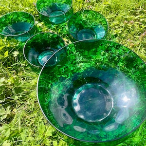 May include: Five green glass bowls of varying sizes, arranged on a bed of green grass. The bowls are clear and have a slight iridescent sheen.