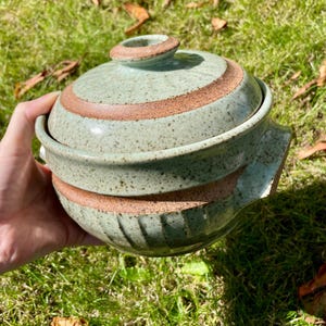 Op de afbeelding: Een groene keramische pot met een deksel, met een gespikkeld glazuur en bruine strepen. De pot heeft een ronde vorm en een klein handvat aan de zijkant.