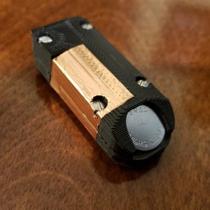 May include: A small, black, cylindrical battery holder with a copper tape wrapped around it. The battery holder has a small, round, silver battery inside with the text "LR44 1.5V 649 CELL" printed on it.