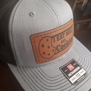 Puede incluir: Una gorra de camionero gris y negra con un parche de cuero marr&oacute;n que dice "I eat milk and cookies". La gorra es ajustable y tiene el logotipo de Richardson 112.