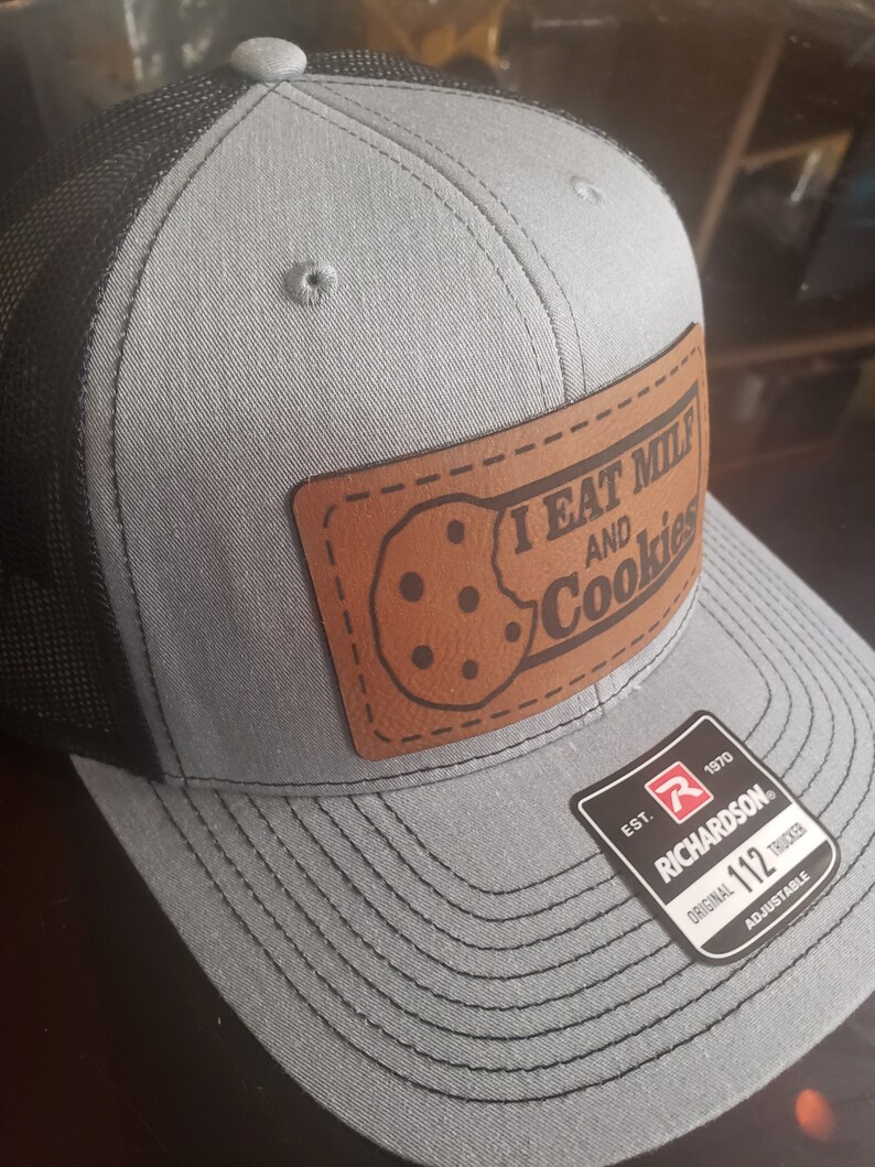 Puede incluir: Una gorra de camionero gris y negra con un parche de cuero marr&oacute;n que dice "I eat milk and cookies". La gorra es ajustable y tiene el logotipo de Richardson 112.