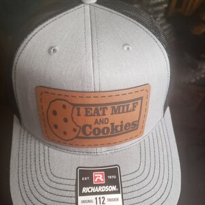 Puede incluir: Una gorra de camionero gris y negra con un parche de cuero marr&oacute;n que dice "I EAT MILF AND Cookies". La gorra tiene una parte trasera de malla negra y un logotipo de Richardson con el texto "EST. 1970 RICHARDSON&reg; ORIGINAL 112 TRUCKER ADJUSTABLE".
