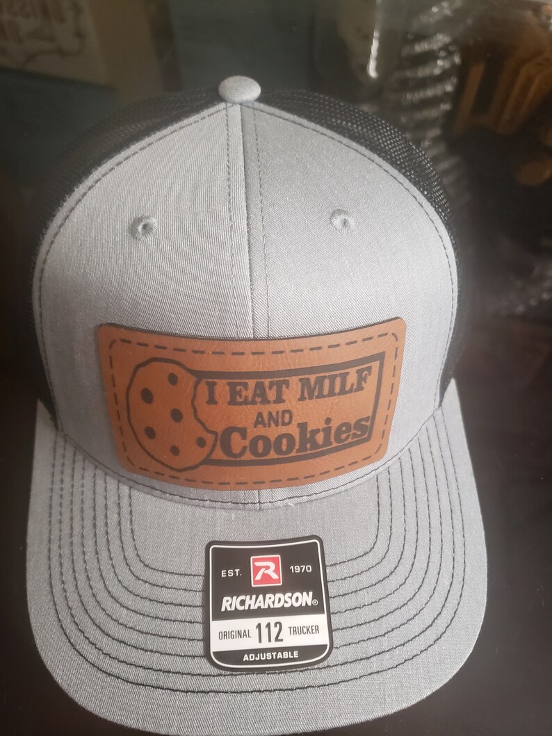 Puede incluir: Una gorra de camionero gris y negra con un parche de cuero marr&oacute;n que dice "I EAT MILF AND Cookies". La gorra tiene una parte trasera de malla negra y un logotipo de Richardson con el texto "EST. 1970 RICHARDSON&reg; ORIGINAL 112 TRUCKER ADJUSTABLE".