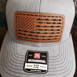 May include: A gray trucker hat with a black mesh back and a brown leather patch. The patch features an American flag design, with fish replacing the stripes. The hat has a curved brim and an adjustable strap. The tag reads "RICHARDSON ORIGINAL 112 TRUCKER ADJUSTABLE".