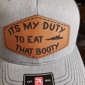 May include: A gray and black trucker hat with a brown leather patch that says "ITS MY DUTY TO EAT THAT BOOTY". The hat has a Richardson logo and the text "EST. 1970 RICHARDSON® ORIGINAL 112 TRUCKER".