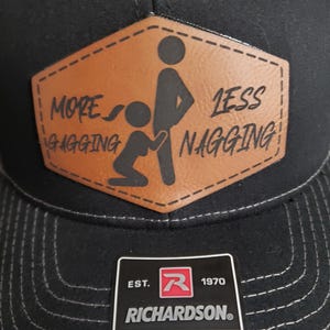 May include: Black trucker hat with a brown leather patch that says "More Gagging Less Nagging" with a stick figure illustration. The hat is adjustable and has a Richardson logo.
