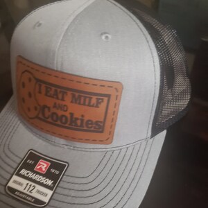 Puede incluir: Una gorra de camionero gris y negra con un parche de cuero marr&oacute;n que dice "I eat milf and cookies". La gorra tiene una parte trasera de malla negra y una parte delantera gris con un logotipo de Richardson.