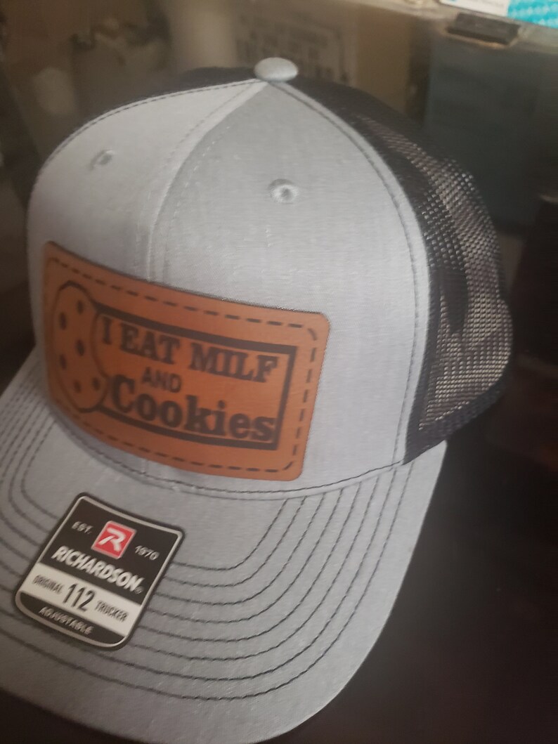 Puede incluir: Una gorra de camionero gris y negra con un parche de cuero marr&oacute;n que dice "I eat milf and cookies". La gorra tiene una parte trasera de malla negra y una parte delantera gris con un logotipo de Richardson.