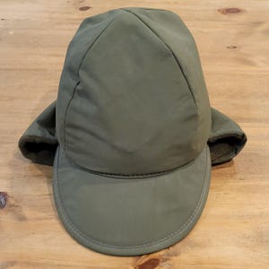 May include: An olive green winter cap with a smooth, water-resistant outer shell and a stiff, rounded brim. The hat features integrated ear flaps lined with a dark olive green fleece material, providing coverage on both sides of the head.