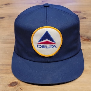 May include: A navy blue baseball cap featuring a circular embroidered patch on the front. The patch has a white background with a yellow border and displays a blue and red triangular logo above the blue text that reads DELTA.