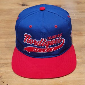 Peut inclure: Une casquette de baseball bleue avec une visière rouge et un bouton rouge sur le dessus. L'avant présente une broderie en rouge et blanc indiquant Quebec Nordiques Hockey, avec Nordiques en écriture cursive et Hockey en lettres capitales.