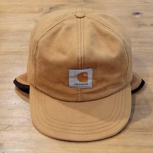 Può includere: Un cappellino da baseball color marrone chiaro con corona strutturata e visiera curva. La parte anteriore presenta una toppa quadrata di colore chiaro con il logo e la scritta Carhartt in marrone. Il cappellino ha cuciture e dettagli neri.
