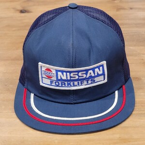 May include: A navy blue baseball cap with a mesh back and a flat brim. The front features a rectangular patch with the text NISSAN FORKLIFTS and a red and white Nissan logo. The brim is accented with a thin white stripe and a red stripe along the edge.