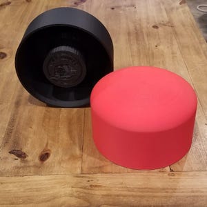 May include: A black, cylindrical object with a circular, recessed design and a red, cylindrical object. The black object has a circular emblem in the center. The red object is a solid, smooth cylinder. Both objects are on a wooden surface.