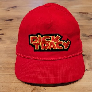 Pode incluir: Um boné de beisebol vermelho brilhante com um emblema bordado proeminente na frente. O emblema exibe o texto "DICK TRACY" em letras amarelas em negrito com contorno vermelho, sobre um fundo preto. Um cordão trançado vermelho decora a aba.