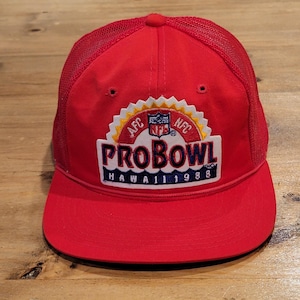 May include: A bright red trucker hat featuring a mesh back and a flat brim. The front panel displays a white embroidered patch with the text PRO BOWL HAWAII 1988, an NFL logo, and AFC and NFC lettering in red, blue, and yellow colors.