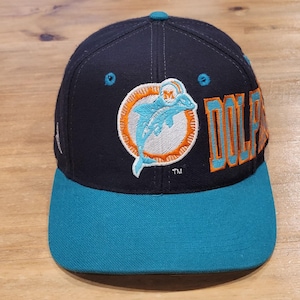 Op de afbeelding: Een marineblauwe honkbalpet met een blauwgroene klep en oogjes. De voorkant is voorzien van een geborduurd Miami Dolphins-logo met een lichtblauwe dolfijn in een oranje cirkel. Het woord DOLPHINS is in oranje en lichtblauw aan de zijkant geborduurd.