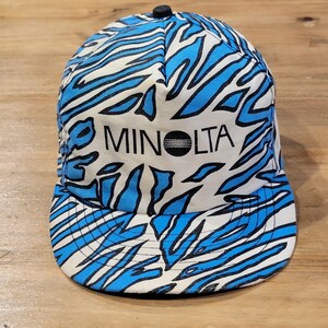 Pode incluir: Um boné de beisebol com base branca e um padrão animal azul e preto semelhante ao de uma zebra. O painel frontal exibe a palavra MINOLTA em letras maiúsculas pretas com um gráfico circular substituindo a letra O. O boné tem um botão preto.