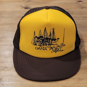 Może przedstawiać: Czapka typu trucker z jaskrawożółtym panelem przednim oraz ciemnobrązową siateczką i daszkiem. Z przodu znajduje się czarna graficzna ilustracja żaglówek w porcie z nadrukowanym poniżej czarnym napisem DANA POINT California.