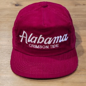 Può includere: Un berretto da baseball in velluto a coste rosso scuro con la parola Alabama in un carattere corsivo bianco, con le parole CRIMSON TIDE stampate in lettere maiuscole bianche direttamente sotto il corsivo sul pannello frontale della corona.
