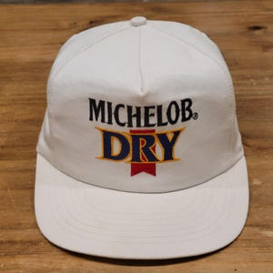May include: A white baseball cap featuring the embroidered text MICHELOB in black above the word DRY in blue with a yellow outline and a red vertical banner graphic underneath. The cap has a structured crown and a flat brim with stitched detailing.