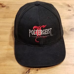Może przedstawiać: Czarna czapka z daszkiem z wyhaftowanym logo z przodu. Wzór zawiera biały napis Poltergeist oraz czerwony napis The Legacy, ze stylizowaną czerwoną literą P wkomponowaną w tło białego napisu.