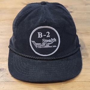 May include: A black corduroy baseball cap featuring a circular patch on the front. The patch displays the text B-2 Stealth in white lettering above a white embroidered graphic of a B-2 bomber aircraft. A black braided cord trim sits above the brim.