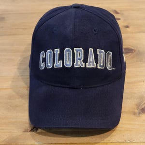 May include: A navy blue baseball cap featuring the word COLORADO embroidered in light blue and white block lettering across the front crown. The cap has a structured six-panel design with a curved brim and a small trademark symbol.