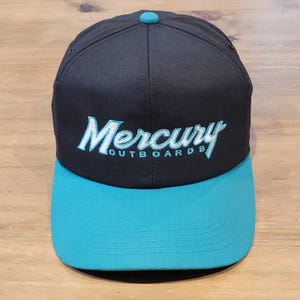 May include: A black baseball cap with a teal brim and a teal button on top. The front features the embroidered text Mercury Outboards in a light blue script font with white outlining, centered on the crown of the structured six-panel hat.