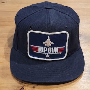 May include: A navy blue baseball cap featuring a rectangular patch on the front. The patch displays a white fighter jet silhouette above the text TOP GUN in white, flanked by red wings and a white star centered below the text on a blue background.