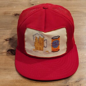 May include: A bright red trucker hat featuring a mesh back and a flat brim. The front panel displays a cream-colored square patch with a cross-stitched design of a yellow beer mug with foam and an orange beverage can with blue and black details.
