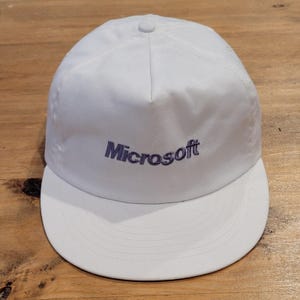 May include: A white baseball cap featuring a five-panel construction with a structured front crown and a flat brim. The word Microsoft is embroidered in a dark purple thread across the center of the front panel in a classic sans-serif font.